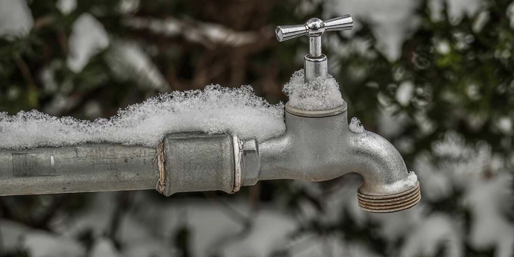 Snow-covered outdoor faucet and water pipe showing winter conditions that can lead to frozen plumbing lines.