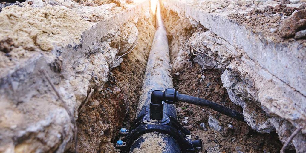 A long plumbing pipe laid in a deep excavated dirt trench beneath concrete, showcasing underground sewer and water line excavation.