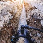 A long plumbing pipe laid in a deep excavated dirt trench beneath concrete, showcasing underground sewer and water line excavation.
