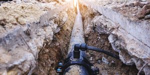 A long plumbing pipe laid in a deep excavated dirt trench beneath concrete, showcasing underground sewer and water line excavation.
