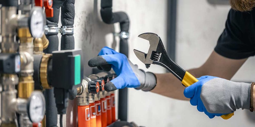 Plumber holding an adjustable wrench while inspecting pipe valves as part of an essential plumbing maintenance guide for homeowners.