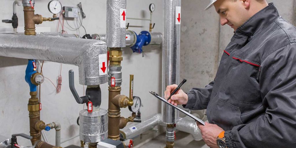 A professional technician inspects insulated pipes and pressure gauges with a checklist for home plumbing maintenance.