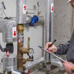 A professional technician inspects insulated pipes and pressure gauges with a checklist for home plumbing maintenance.