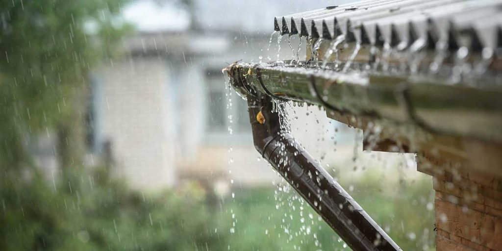 Heavy rain overflows a house roof gutter and downspout, illustrating potential storm impact on home drainage.