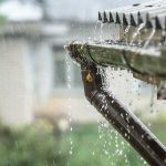 Heavy rain overflows a house roof gutter and downspout, illustrating potential storm impact on home drainage.