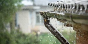 Heavy rain overflows a house roof gutter and downspout, illustrating potential storm impact on home drainage.